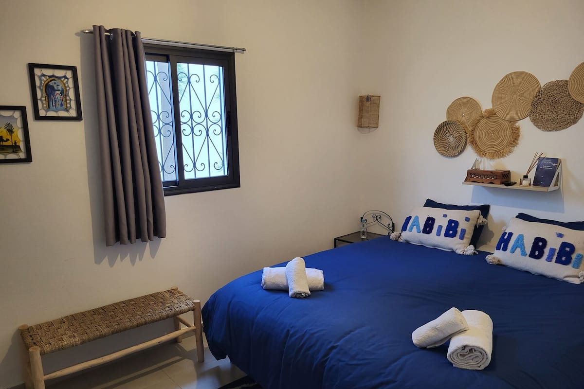 A bedroom with a large bed, a bench, a window with curtains, and wall decorations.