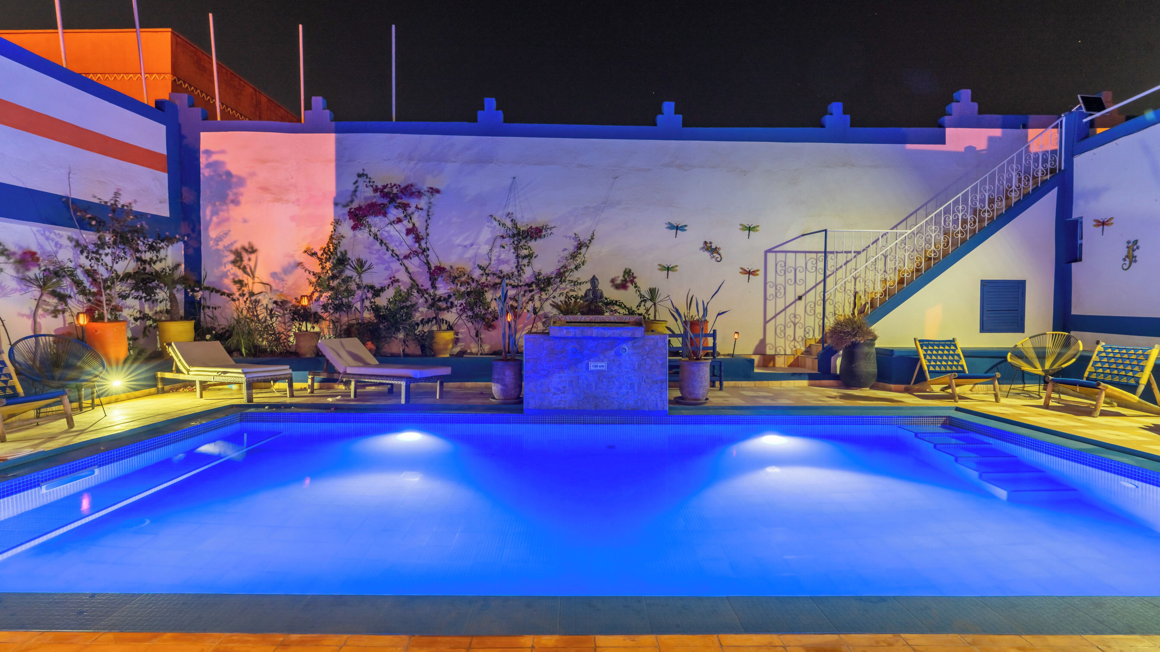 Piscina con sillones de descanso y plantas en macetas, rodeada por un muro blanco con detalles decorativos.