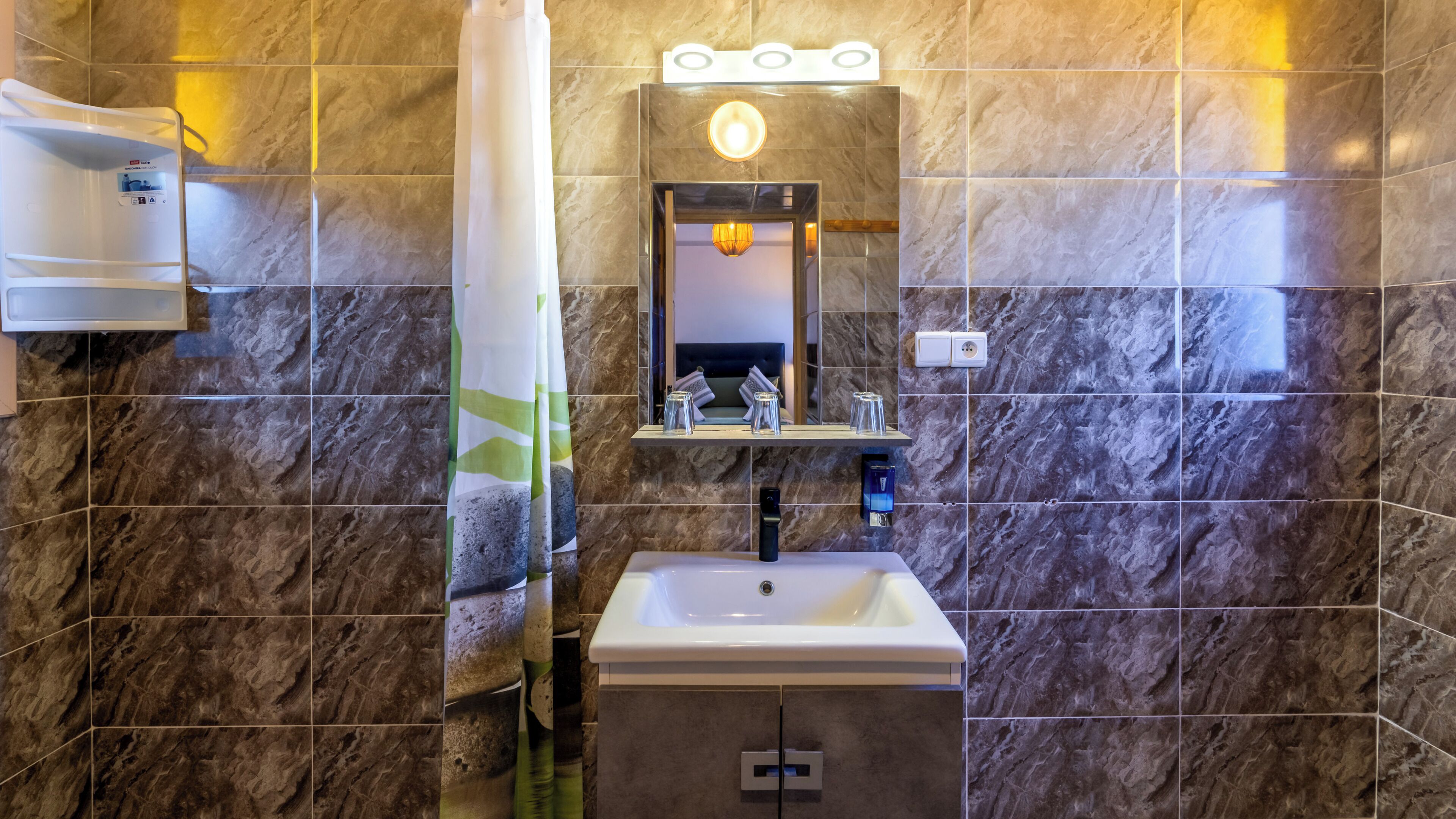 Een badkamer met een betegelde muur van marmer, een wit wastafelmeubel, een spiegel met verlichting erboven en een handdoekrek.