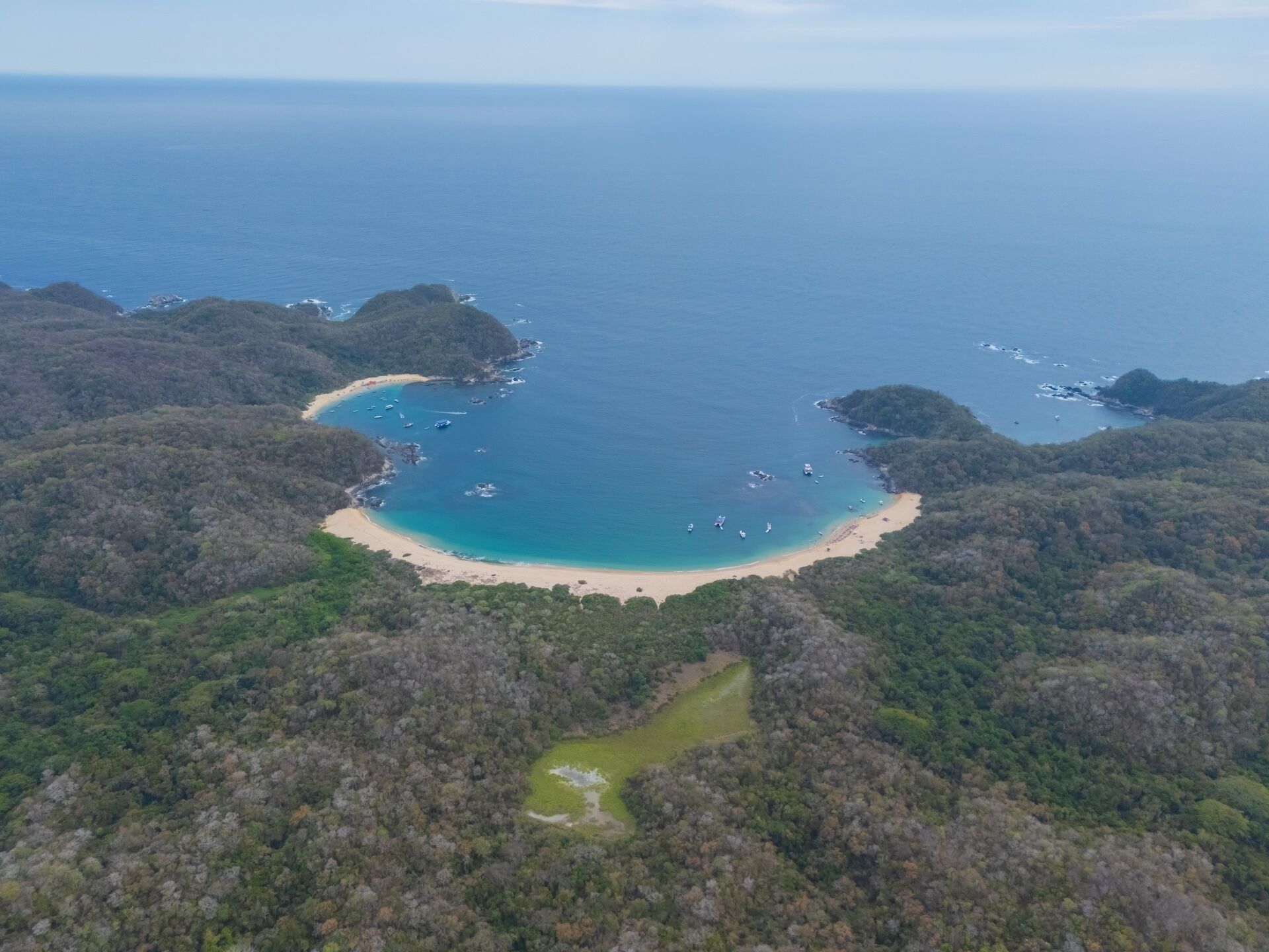 An aerial view of a coastal area with a beach, boats, and forested hills.