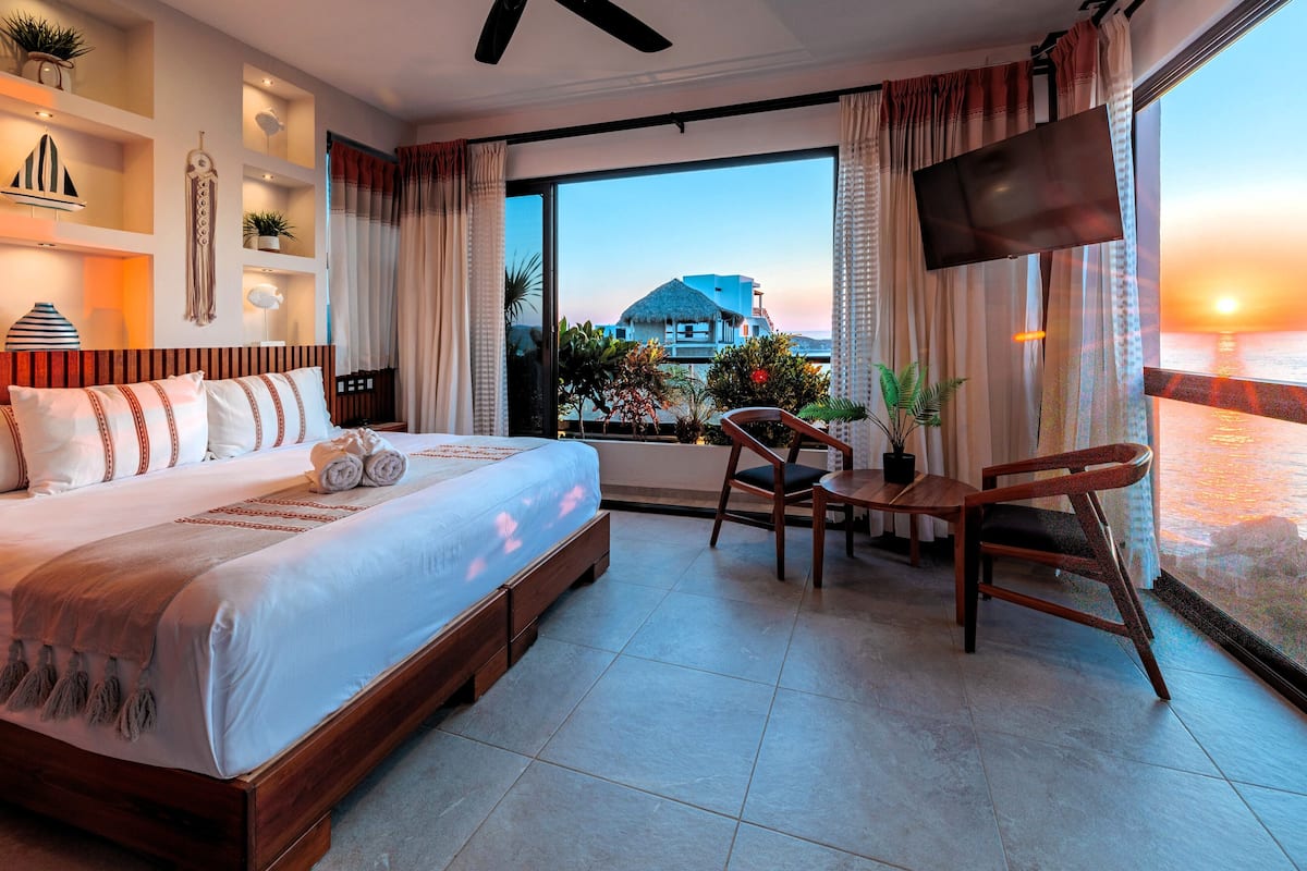A hotel room with a bed, a TV, a dining area, and a view of the sunset over the ocean.