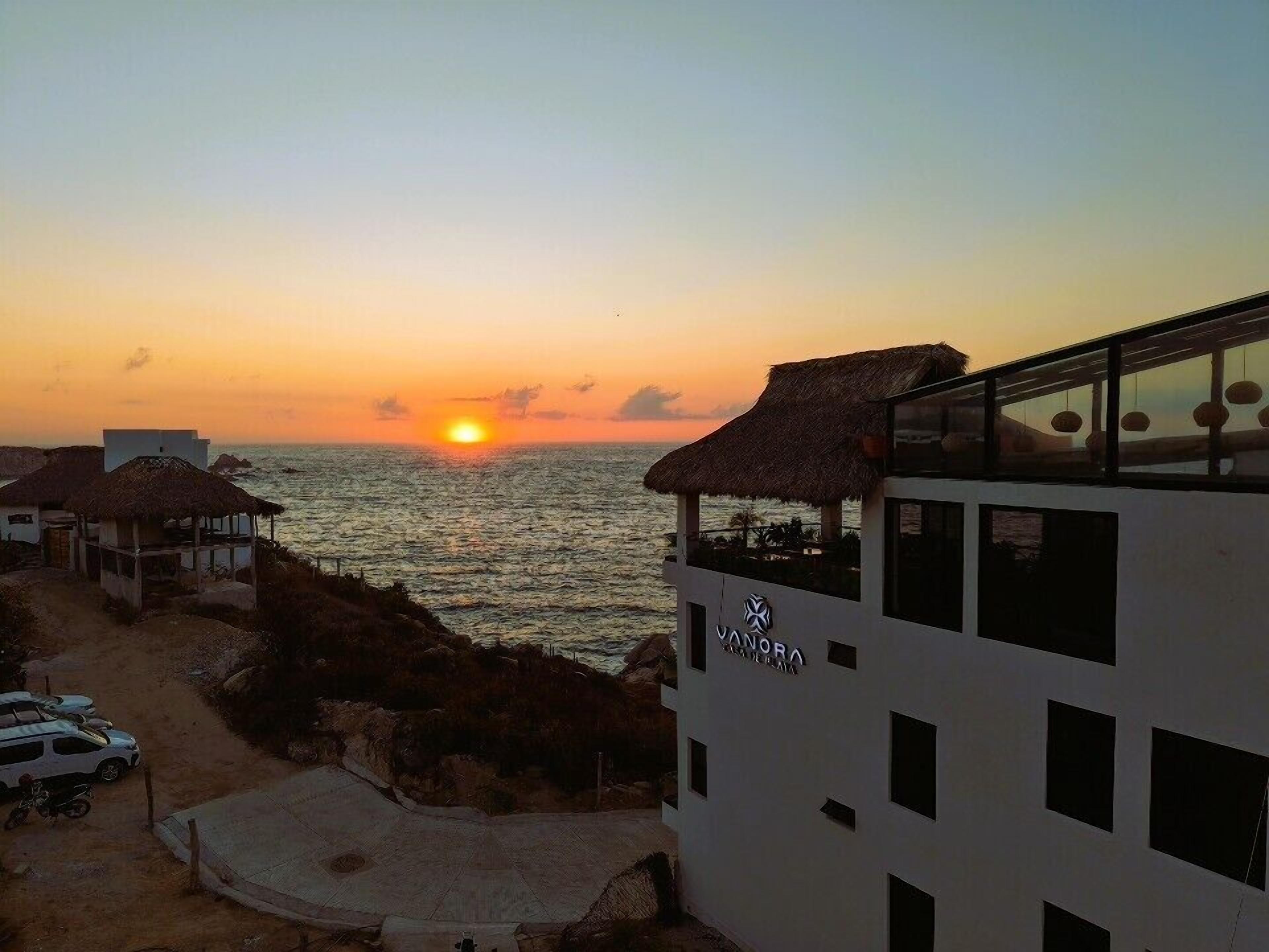 A beachfront hotel with a thatched-roof structure, a pool area, and a view of the sunset over the ocean.