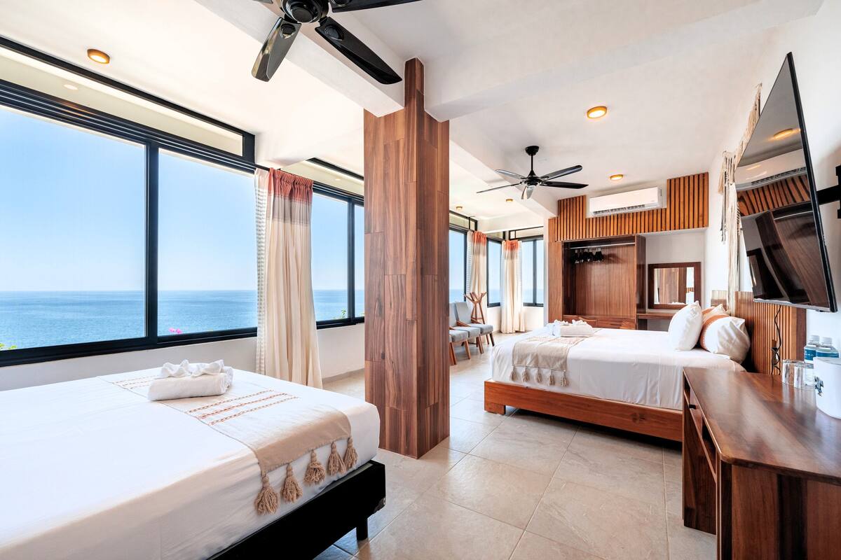 A hotel room with two beds, a TV, a desk, and a large window with a view of the ocean.