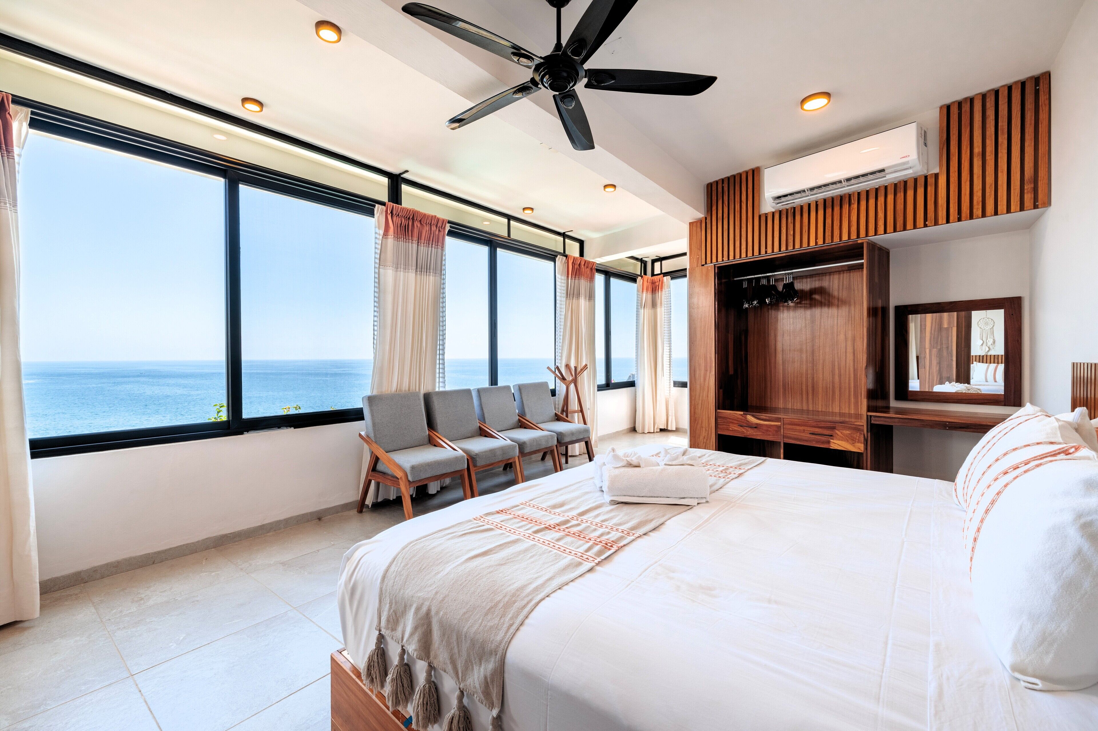 Un dormitorio amplio con una cama grande, un ventilador de techo y vistas al mar a través de amplias ventanas.