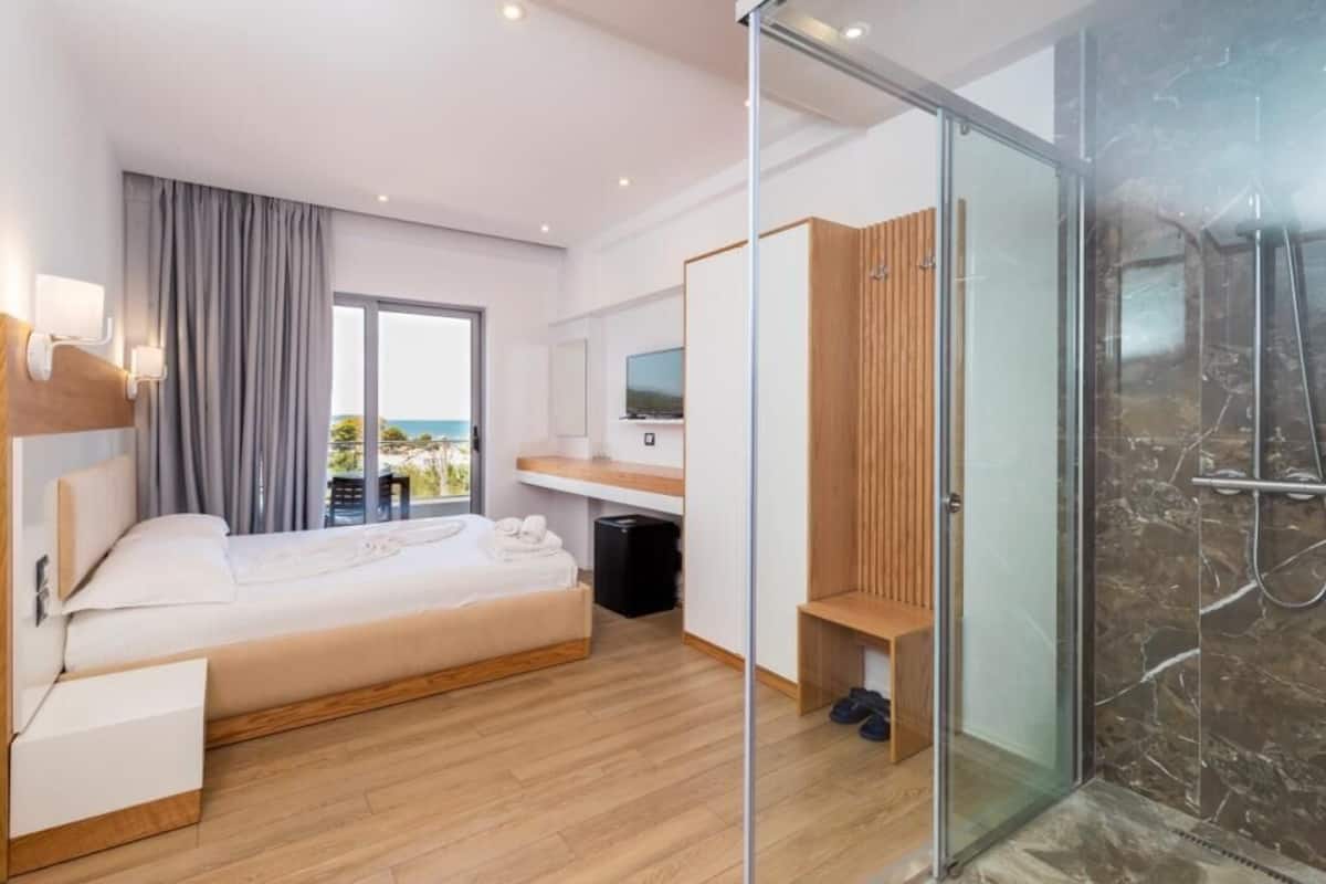A modern hotel room with a bed, a desk, a TV, a shower area, and a view of the outdoors.