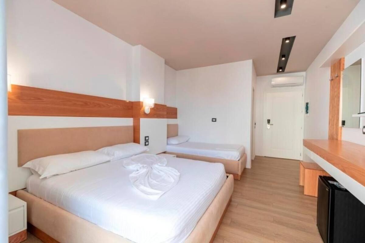 A hotel room with two beds, a wooden headboard, a bedside table, a mirror, and a small kitchenette.