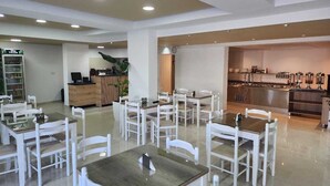Un amplio comedor con sillas y mesas blancas, una recepción y una zona de cocina con mesas tipo bufé.