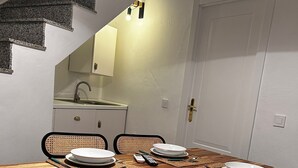 On aperçoit un coin repas avec une table en bois dressée pour quatre personnes, entourée de chaises. À l’arrière-plan, on distingue une cuisine équipée d’un évier et de placards.