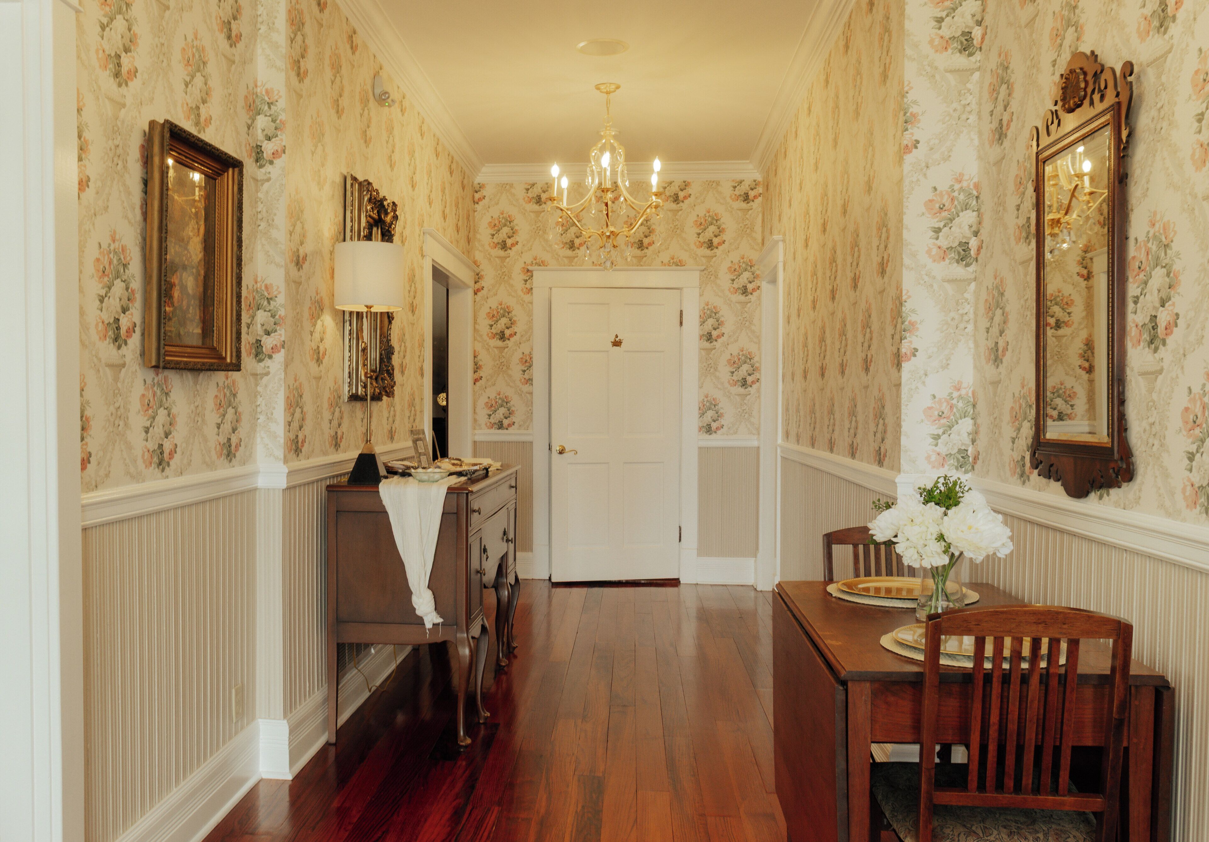 A hallway with floral wallpaper, a chandelier, a white door, and a dining table with chairs.