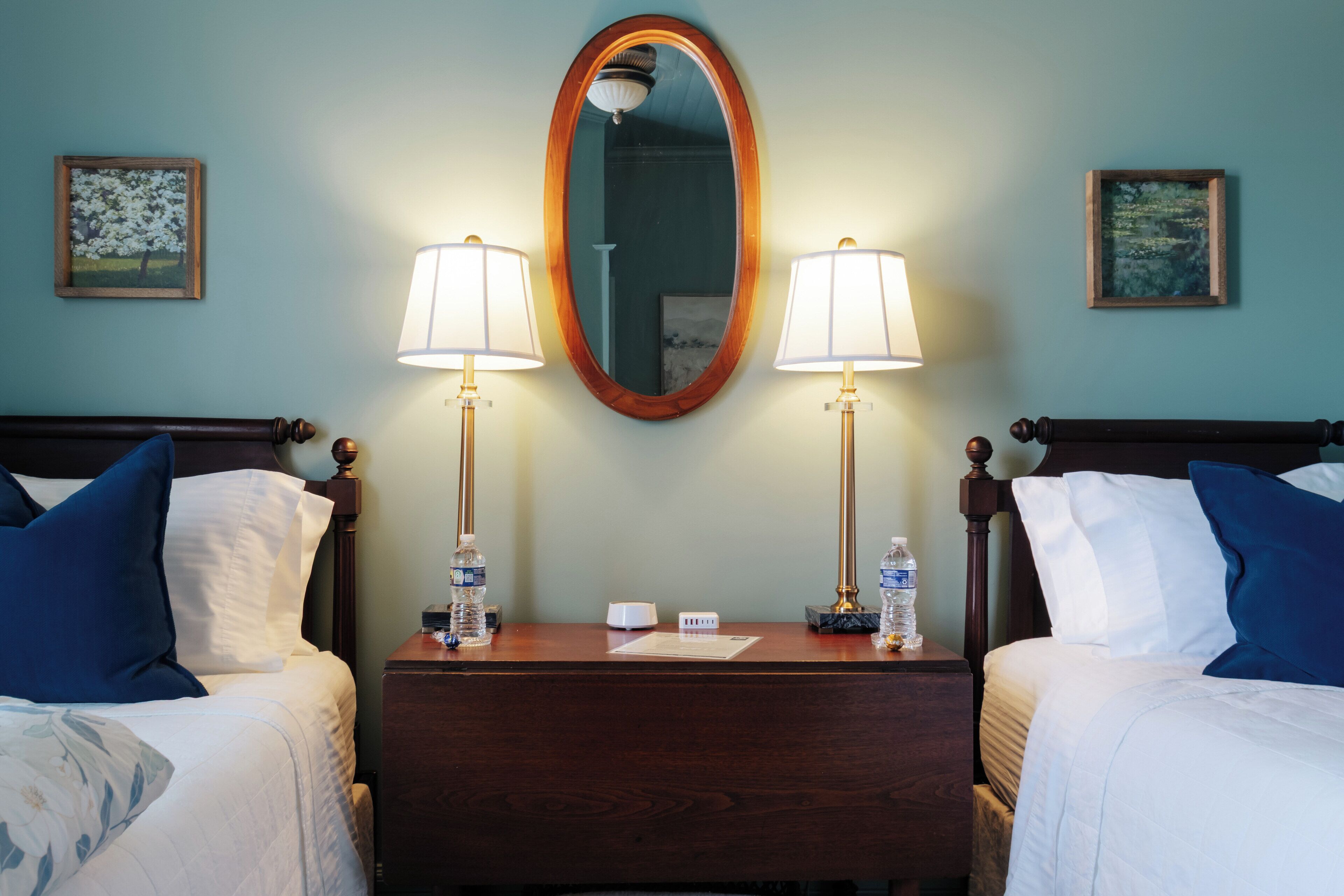 A hotel room with two beds, a wooden nightstand, two bedside lamps, two framed pictures on the wall, and a mirror above the nightstand.