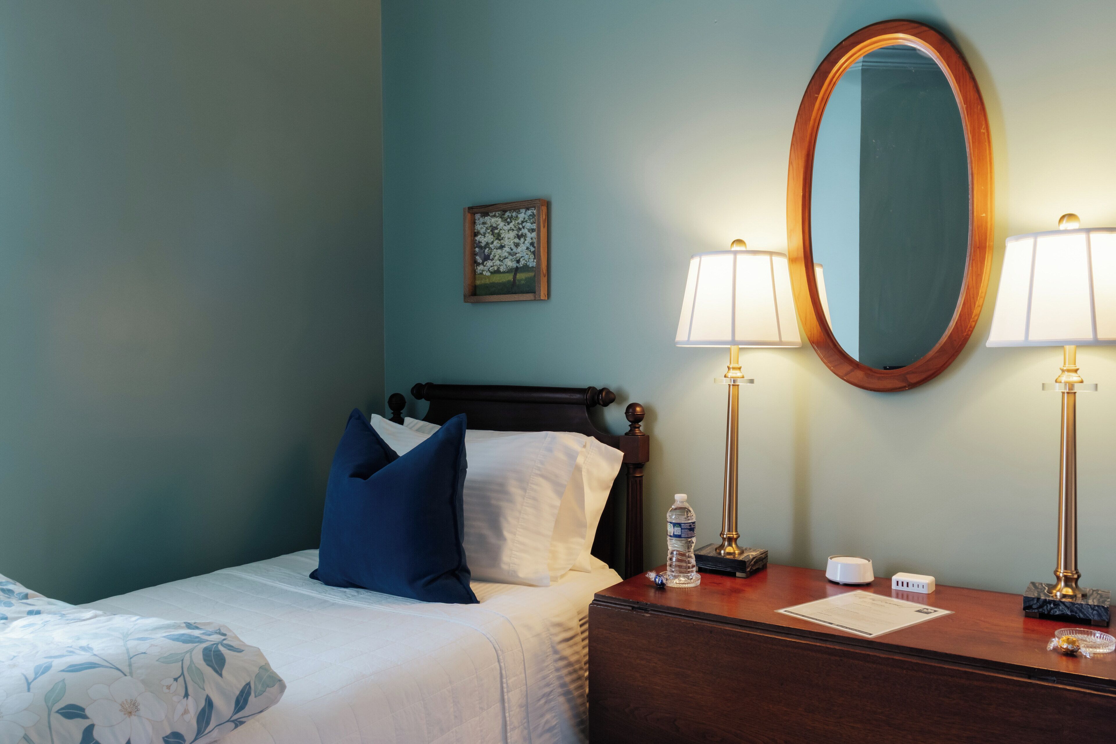 A neatly made bed with a floral-patterned pillow, a wooden headboard, and a nightstand with a lamp, a water bottle, and a small clock.