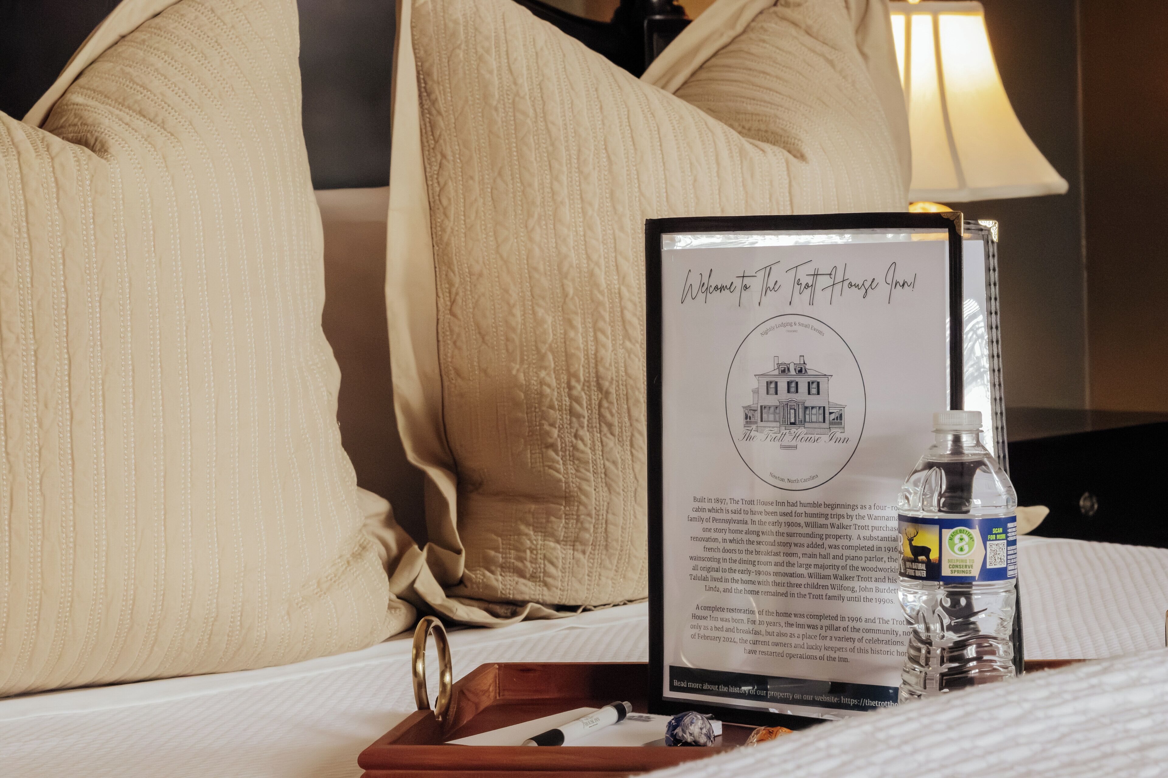 A framed welcome sign for The Trott House Inn, a bottle of water, and a wooden tray on a bed.