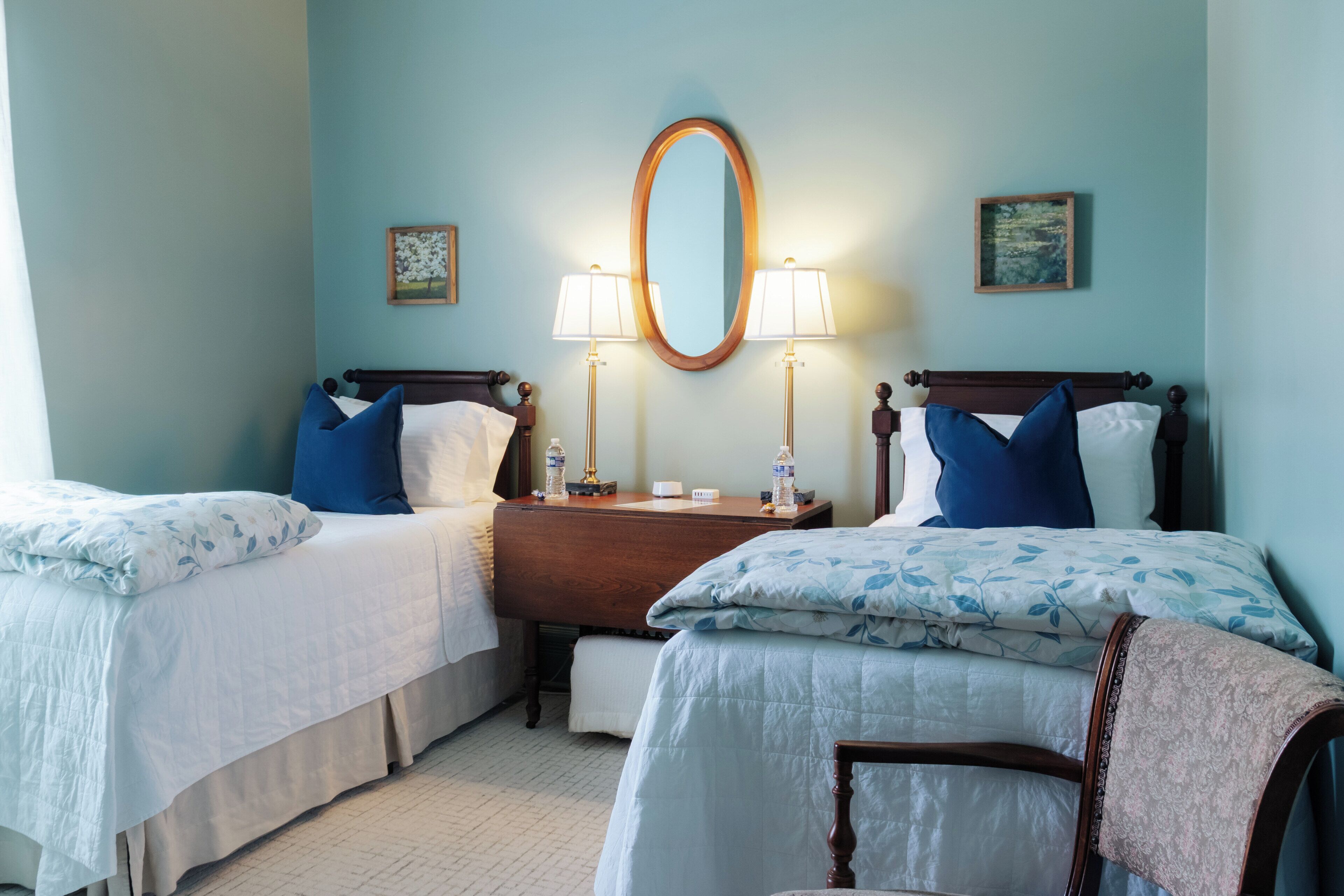Une chambre d’hôtel avec deux lits, une commode en bois, un miroir et deux lampes fixées au mur.