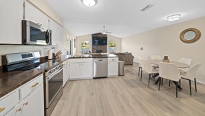 A modern kitchen with white cabinets, stainless steel appliances, and a dining area with white chairs and a round table. - Centrally Located, Serene Luxury Retreat (Brandon)