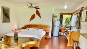 A bedroom with a wooden bed, a dining table with stools, a ceiling fan, and a kitchenette with a refrigerator. - The Fruit Tree Garden Bromeliad Suite (Arenal)