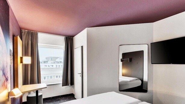 A hotel room with a bed, a bedside table, a mirror, a TV, and a window with curtains. - B&B Hotel Bremen-altstadt (Bremen)