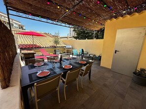 An outdoor patio with a table set for dining, surrounded by chairs, under a thatched roof with colorful lights.