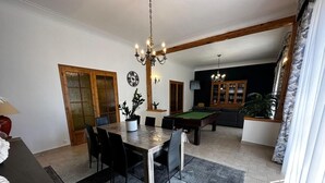 A dining area with a pool table, chairs, and a chandelier. There is a sofa and a cabinet with books in the background.
