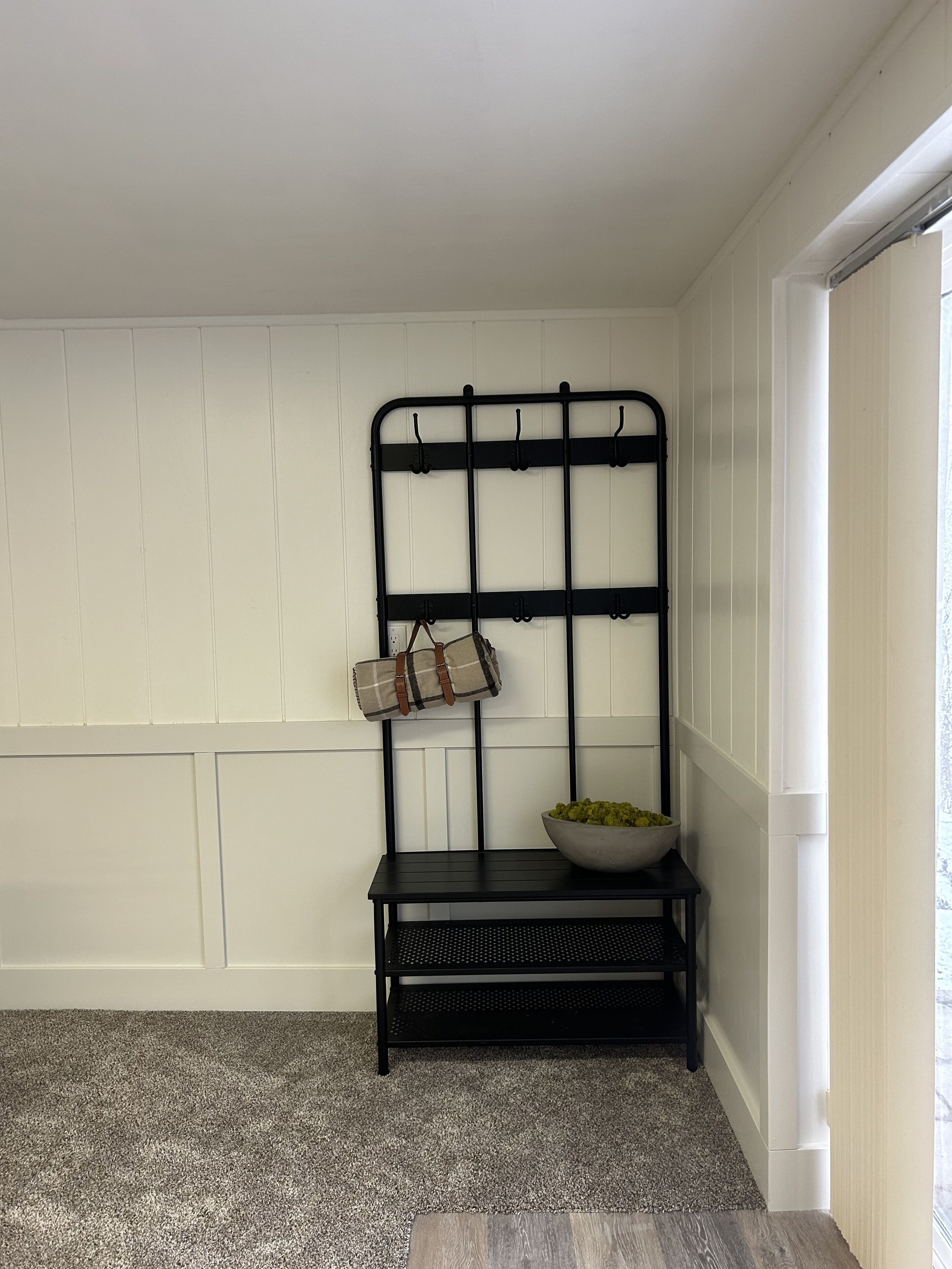 A room with a black metal rack against a white paneled wall, a small black table with a bowl of fruit, and a grey carpet.