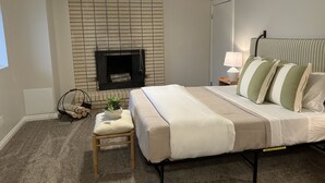 A bedroom with a bed, a bedside table with a lamp, a small stool, a fireplace, and a window. - Cozy Mountain Retreat - Snowbasin, Skiing, Biking, Hiking, Fishing (Morgan)
