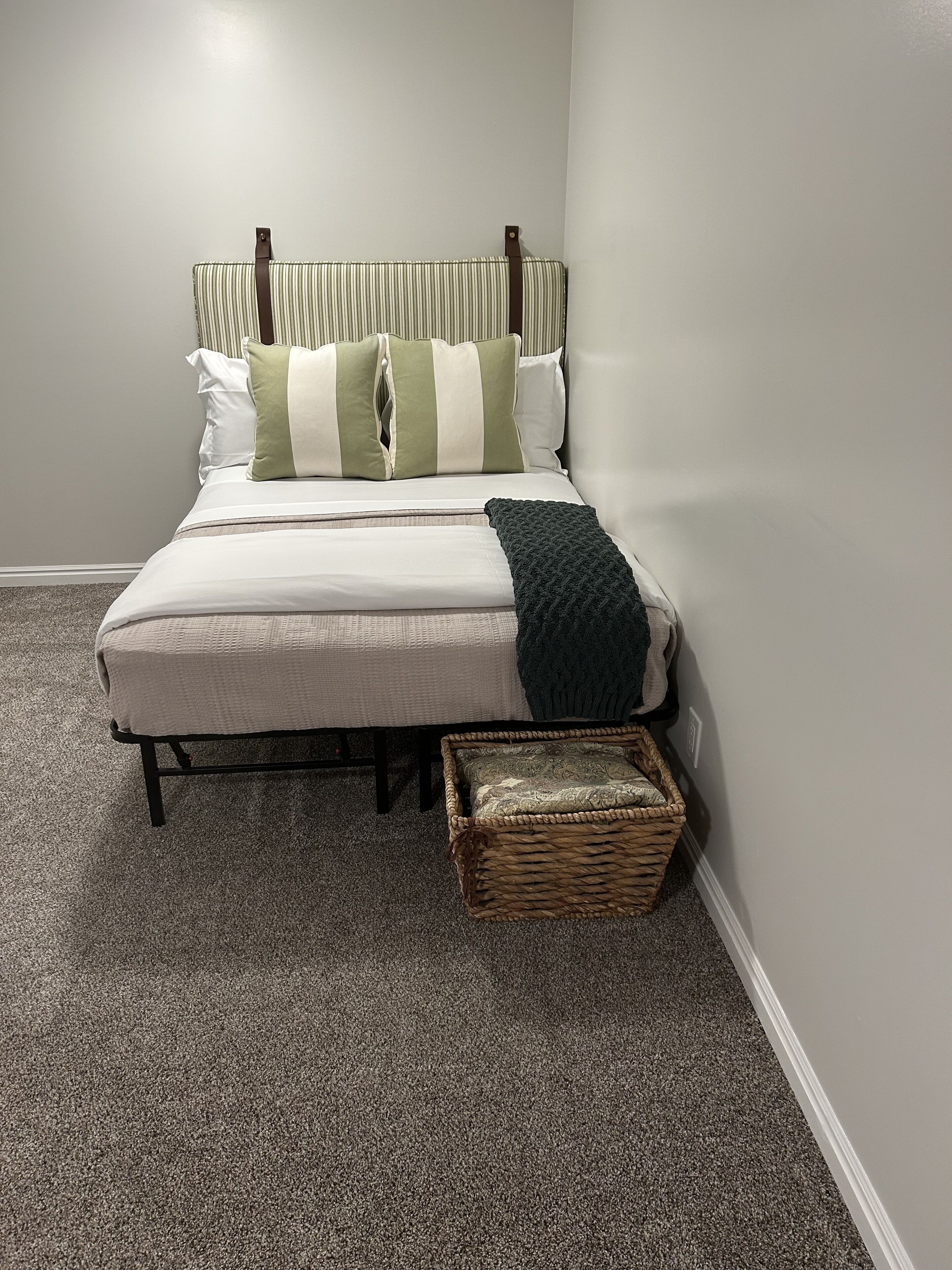 Een net opgemaakt bed met gestreept beddengoed en kussens, een donge dekensprei en een rieten mand aan het voeteneinde.