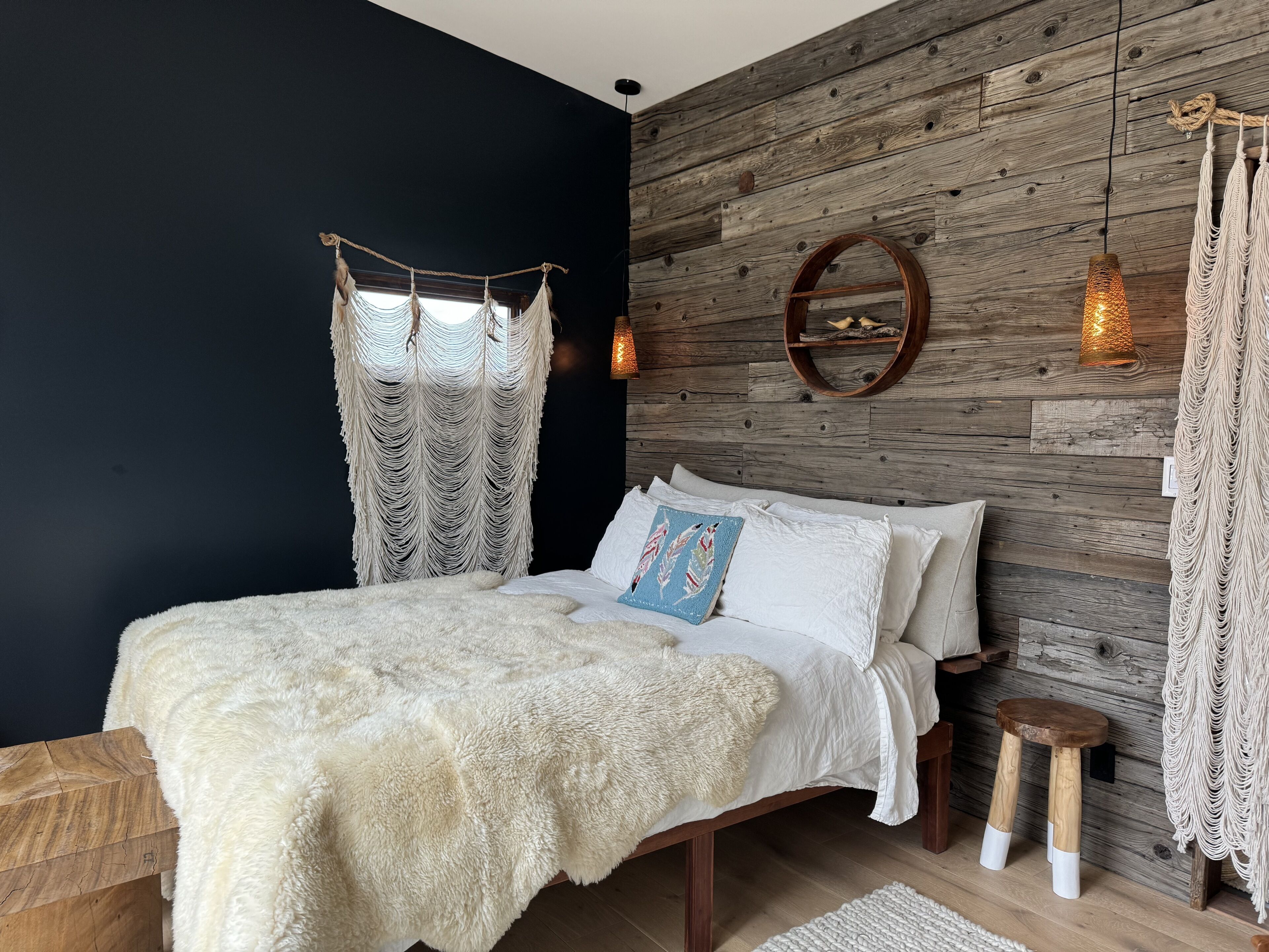 A bedroom with a bed, a wooden wall, a macrame curtain, a round mirror, and a hanging light fixture.