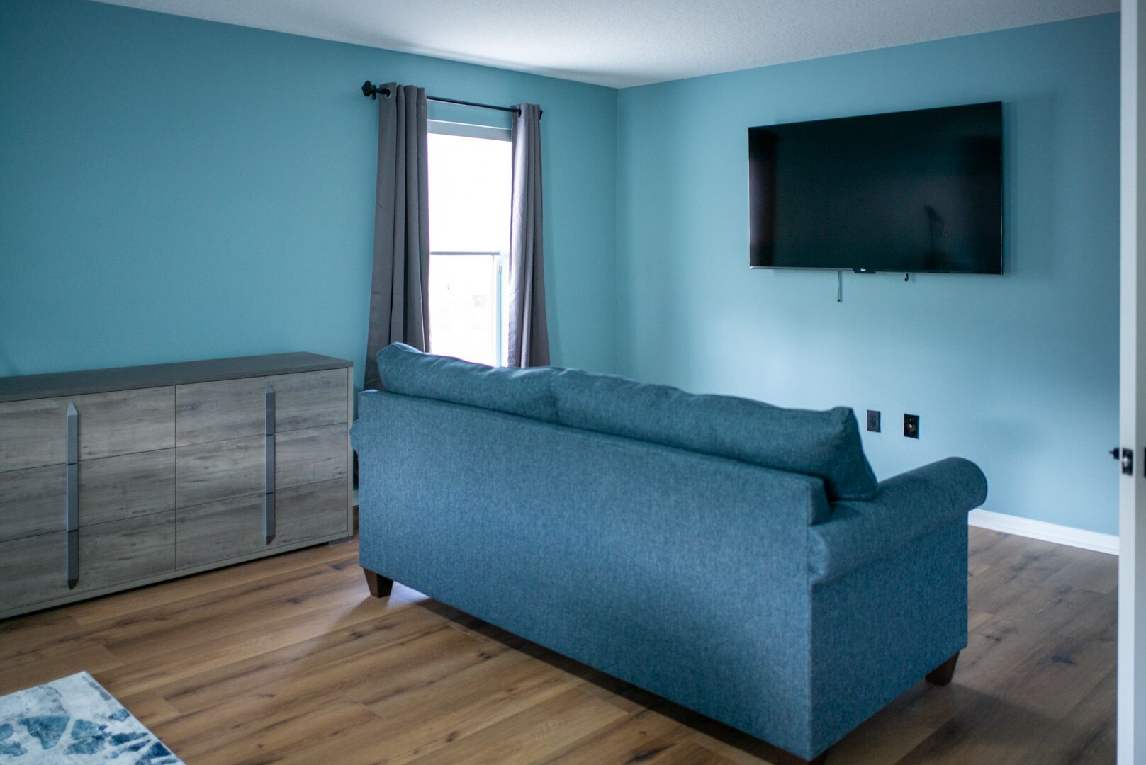 A room with a blue sofa, a wooden cabinet, a TV on the wall, and a window with curtains.