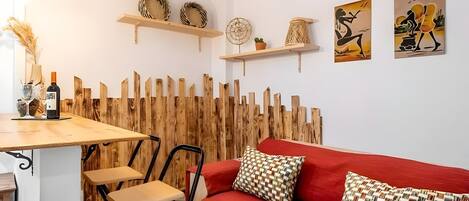 A cozy room with a red sofa, two chairs, a wooden table with a bottle and glass, and decorative items on shelves.