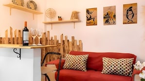Un salon avec un canapé rouge, une table basse en bois et un coin bar avec une bouteille de vin et des verres. Des étagères murales sont ornées d’objets décoratifs et d’œuvres d’art encadrées.