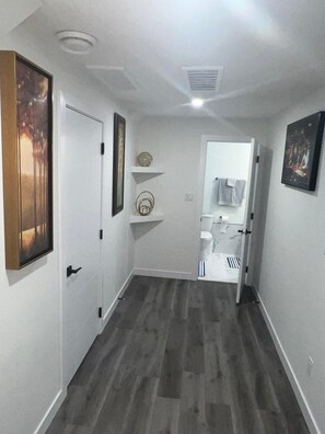 A narrow hallway with a wooden floor, white walls, and a door leading to a bathroom. There are two framed pictures on the walls, a small shelf with decorative items, and a light fixture on the ceiling. - Comfy 2 bedroom stay tucked away in a sleepy neighborhood few mins to the Henday (Edmonton)