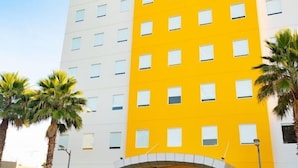 A multi-story hotel building with a yellow facade and a white lower section. There is a white van parked in front with a person standing beside it. - One Irapuato (Irapuato)