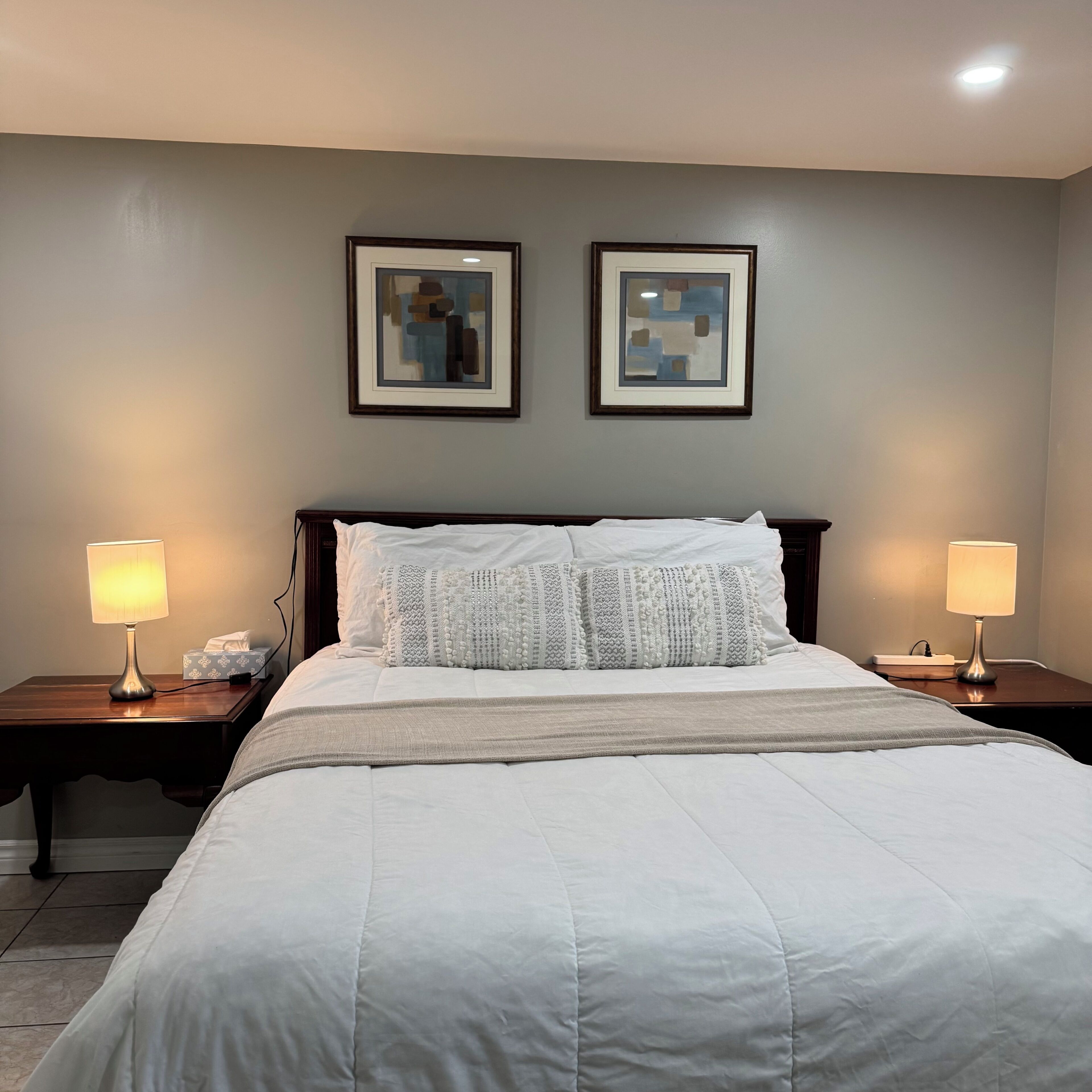 Un lit bien fait, recouvert d’une couette blanche et agrémenté de plusieurs oreillers, est flanqué de deux tables de chevet sur lesquelles reposent des lampes. Le tout est placé devant un mur orné de deux tableaux abstraits encadrés.
