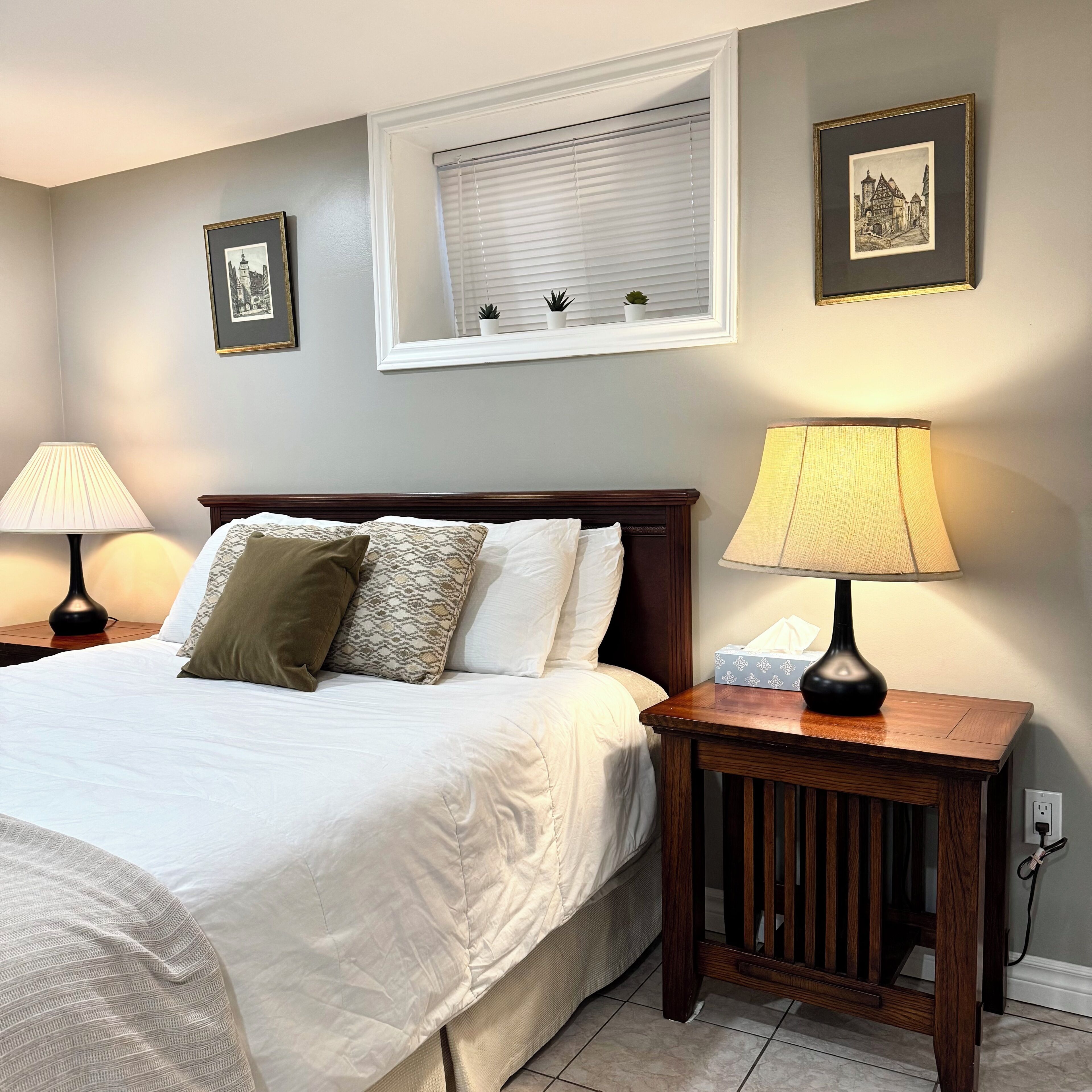 A neatly made bed with pillows, a bedside table with a lamp, and a window with blinds.