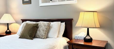 A neatly made bed with pillows, a bedside table with a lamp, and a window with blinds.