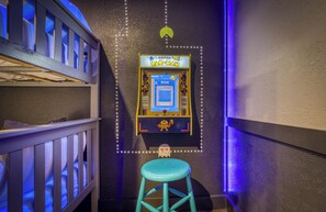 Une borne d’arcade avec le jeu Pac-Man, un tabouret bleu et un lit superposé dans une pièce à la lumière tamisée.