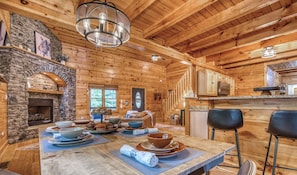 A wooden cabin interior with a stone fireplace, dining table set with bowls and plates, a chandelier, and a kitchen area.