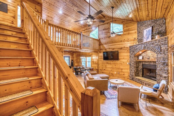 A wooden cabin interior with a staircase, a stone fireplace, a television, and a dining area.