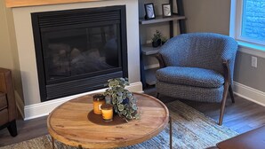 A living room with a fireplace, a flat-screen TV mounted above it, a round coffee table with candles and a plant, a patterned armchair, and a brown leather sofa.