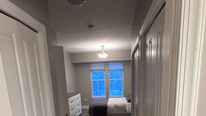 A hallway leading to a bedroom with a bed, dresser, and windows with blinds.