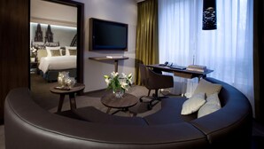 A modern hotel room with a curved seating area, a flat-screen TV, a desk with a chair, and a bed with a view of a cathedral. - Lindner Hotel City Plaza Formerly Renaissance Cologne Hotel (Cologne)
