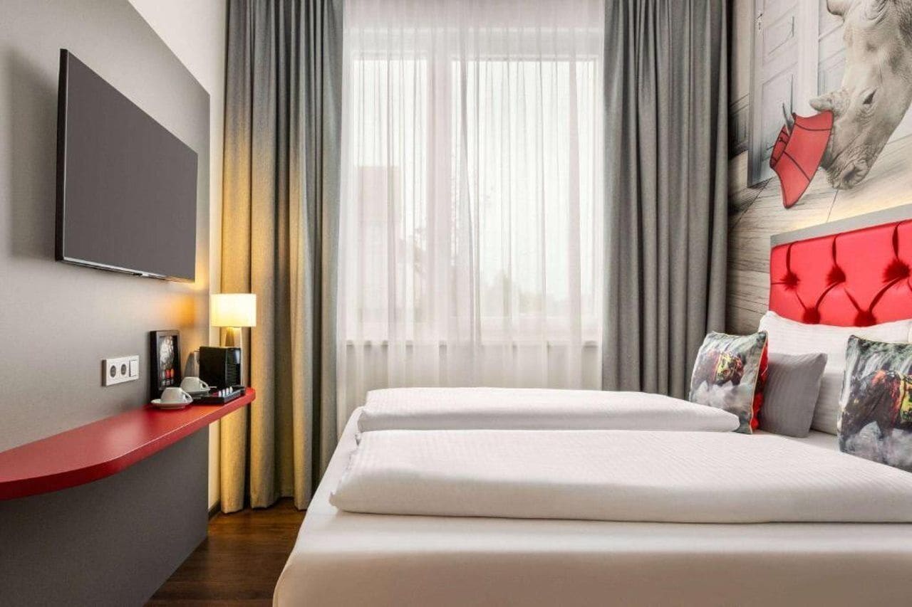 Hotellihuone, jossa on sänky, televisio, punainen työpöytä, lamppu ja ikkuna, jossa on verhot.