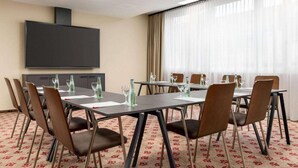 Een vergaderruimte met een flatscreen televisie, meerdere tafels met stoelen en een groot raam met gordijnen.