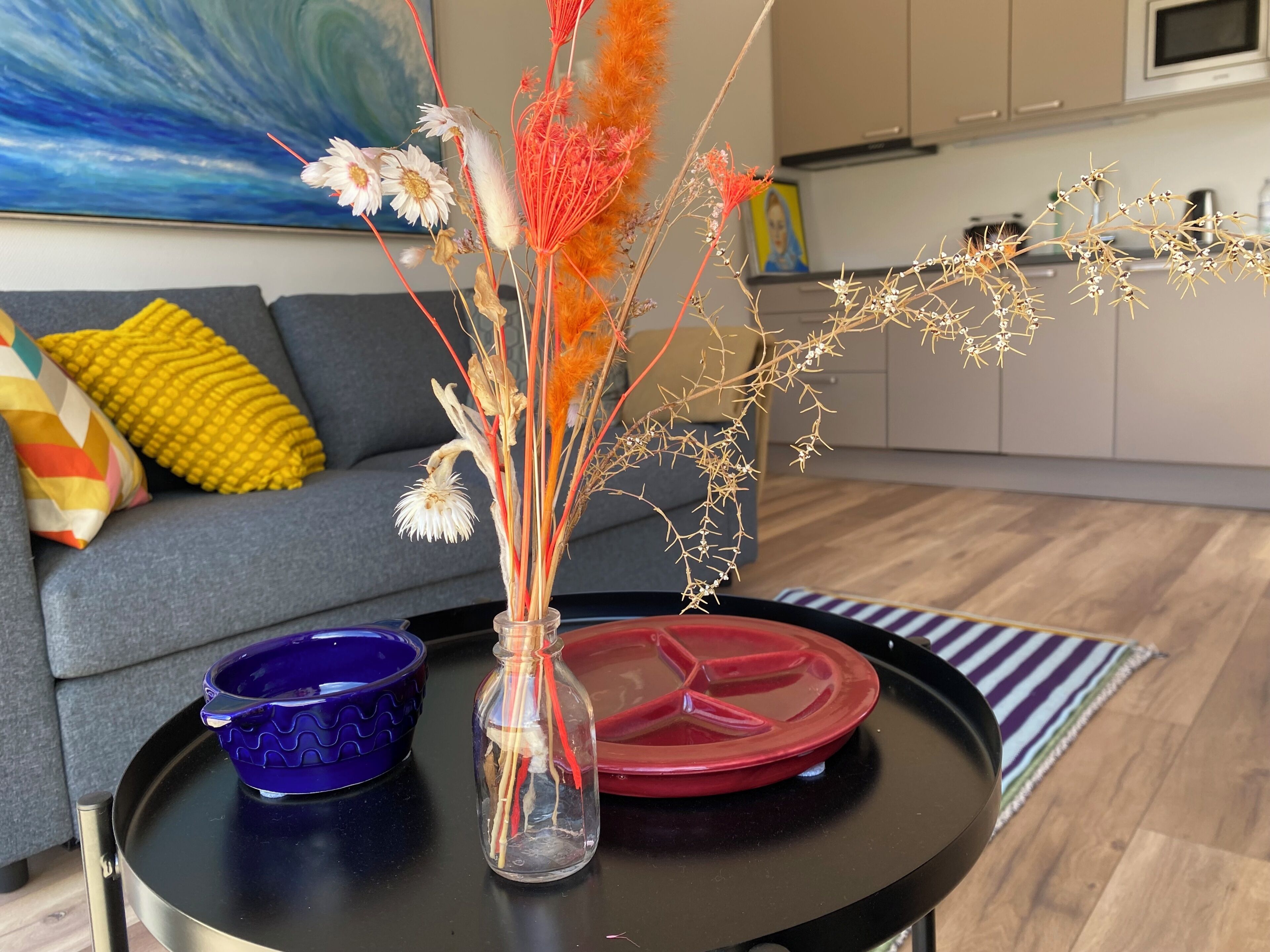 Un salon avec un canapé, une table basse sur laquelle se trouve un vase de fleurs séchées, un bol bleu et une assiette rouge.