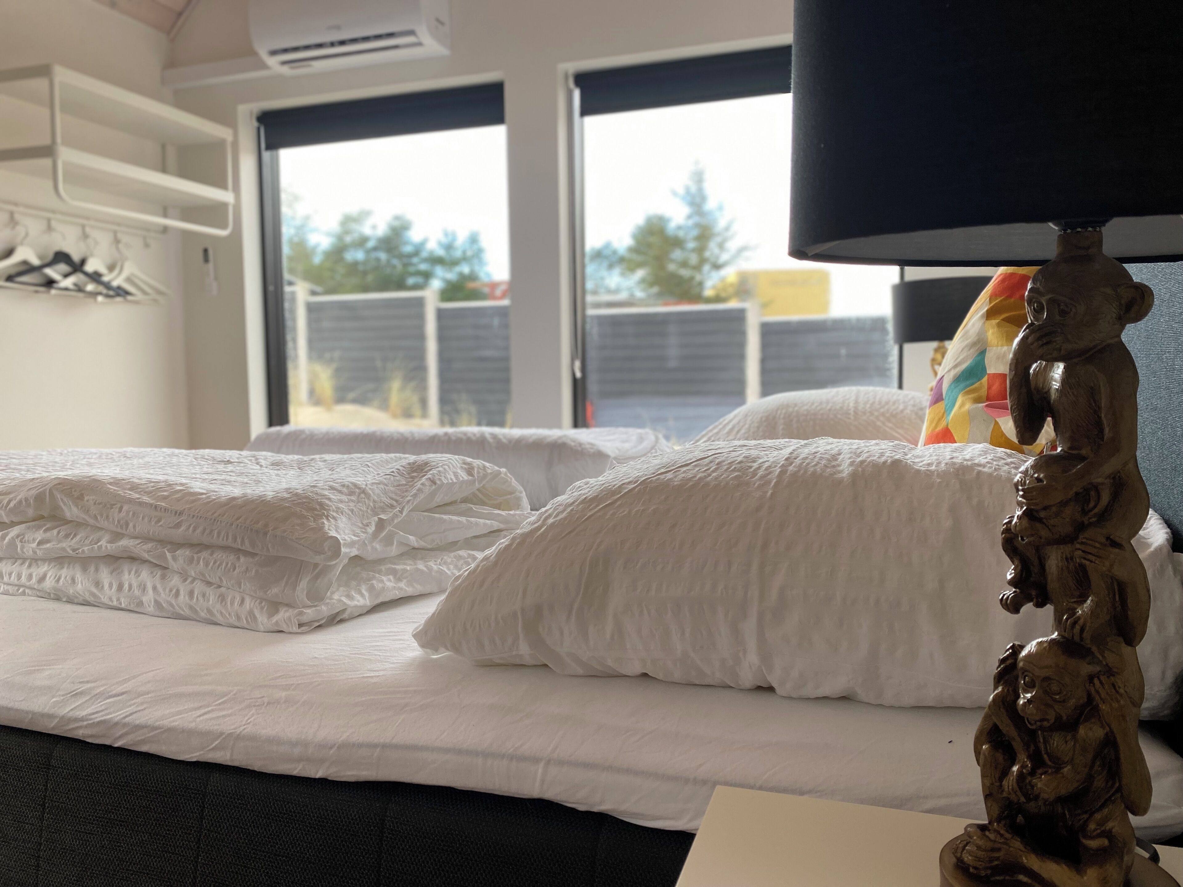 A hotel room with a bed, a wall-mounted shelf with hangers, a window with a view of trees and buildings, and a decorative wooden figure on the bedside table.