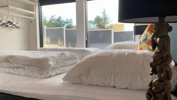 A hotel room with a bed, a wall-mounted shelf with hangers, a window with a view of trees and buildings, and a decorative wooden figure on the bedside table.