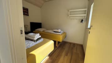 Una habitación pequeña y funcional con dos camas, una mesita de noche, un estante en la pared y suelo de madera.