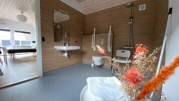 Une salle de bain moderne avec des murs recouverts de panneaux en bois, un lavabo blanc, des toilettes et une douche. On aperçoit un lit dans la pièce adjacente.