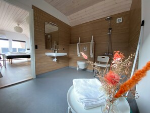 A modern bathroom with wooden panel walls, a white sink, a toilet, and a shower. There is a bed visible in the adjacent room. - Esehytter Luxury Holiday Home near Beach (Norre Nebel)