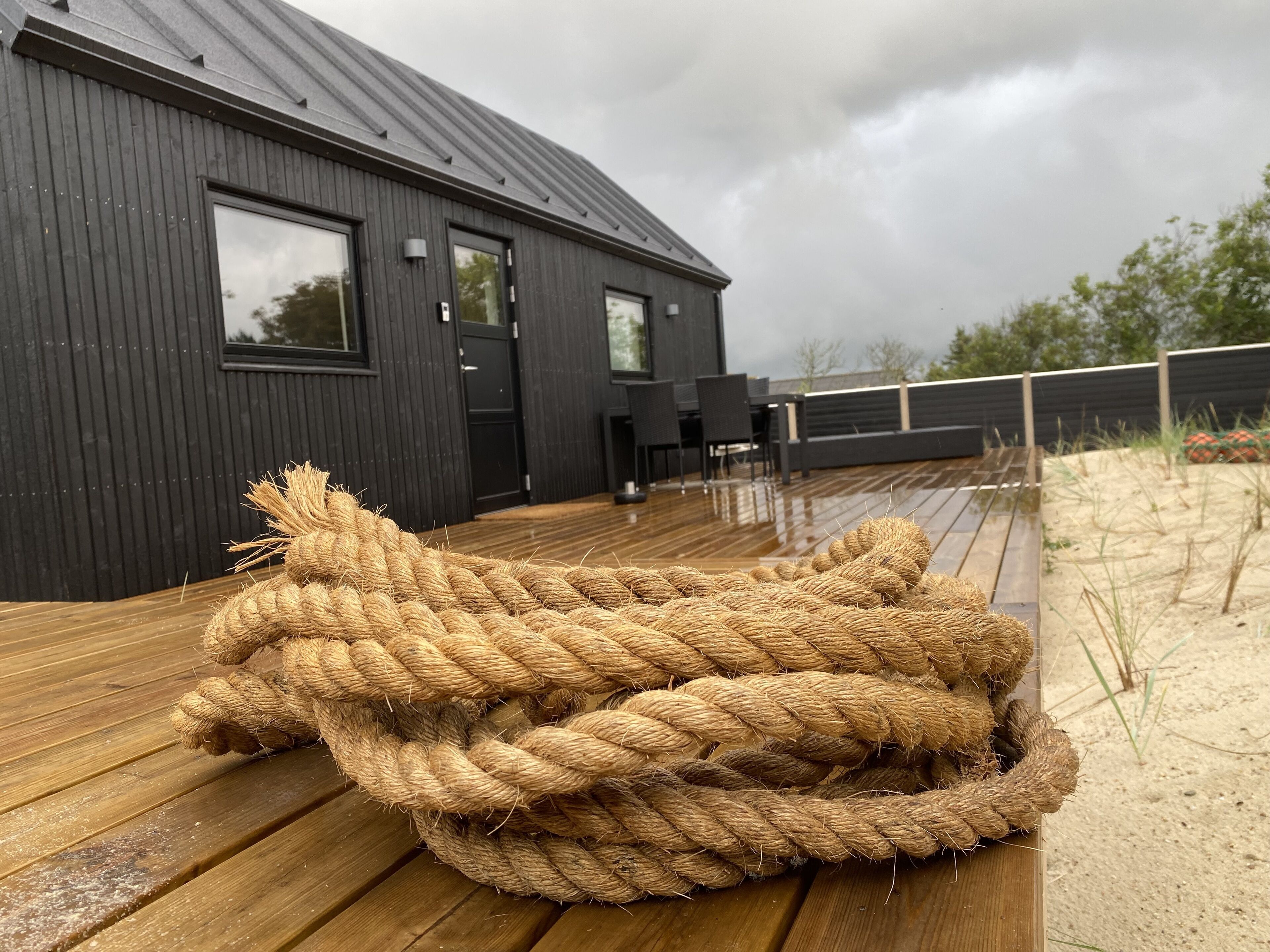 A wooden deck with a large rope bundle in the foreground, a dark wooden building with windows and a door in the background, and a cloudy sky.