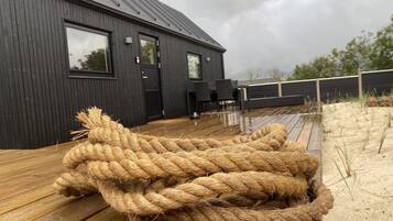 A wooden deck with a large rope bundle in the foreground, a dark wooden building with windows and a door in the background, and a cloudy sky.